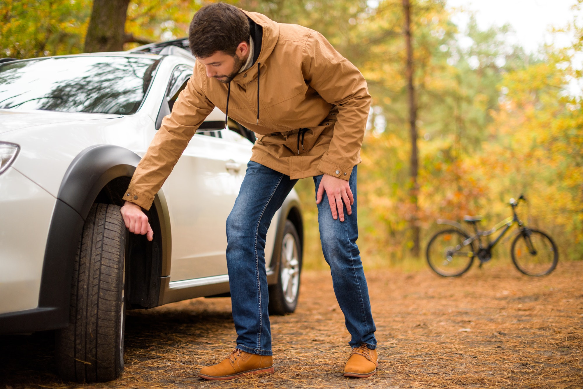 Young bearded man checking car tyre in autumn forest