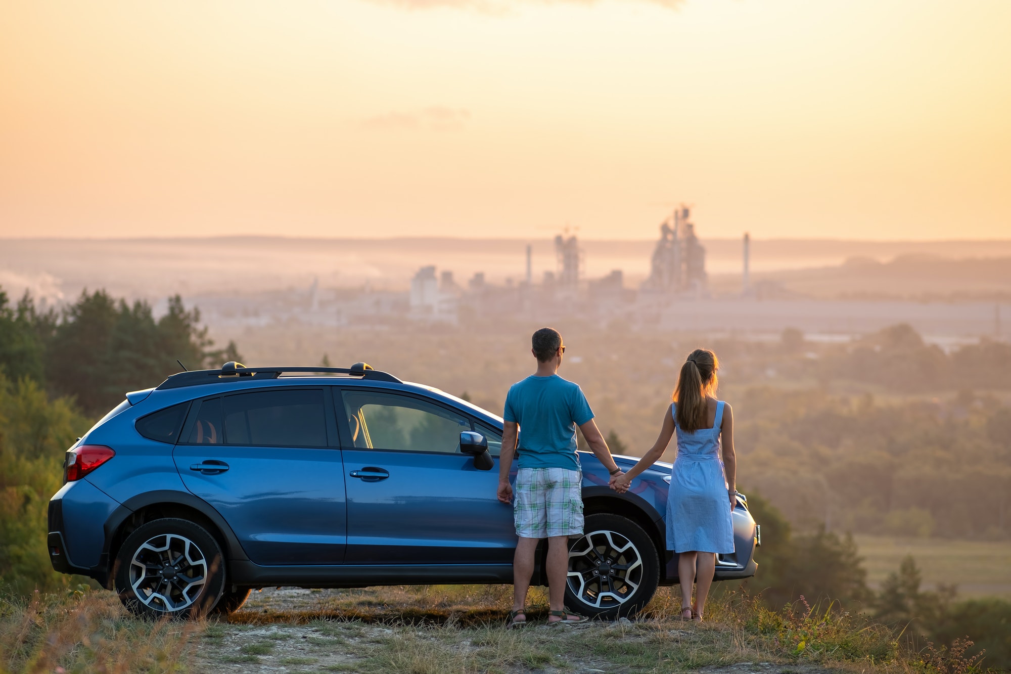 Happy couple relaxing beside their SUV car during honeymoon road trip at sunset. Young man and woman