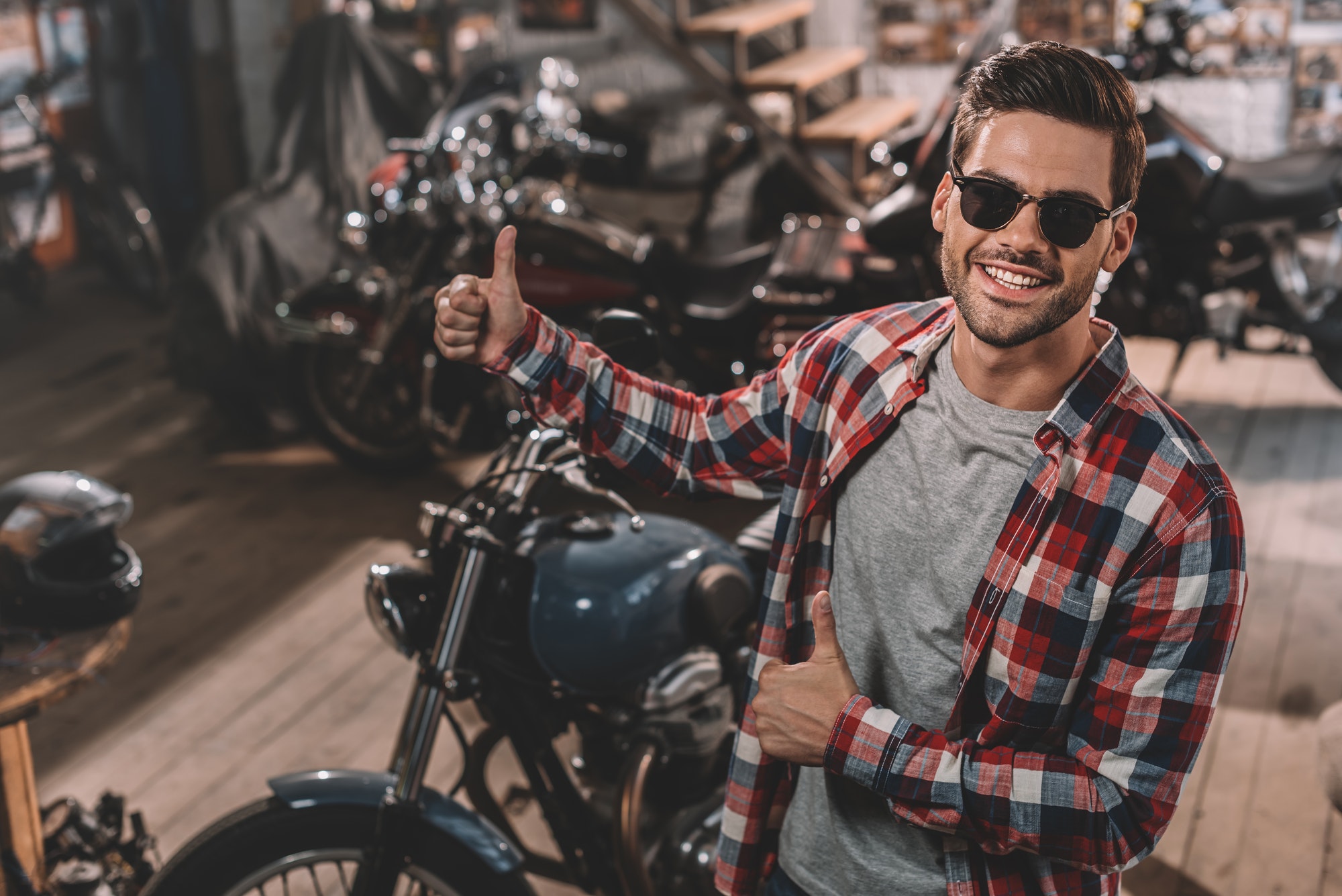 happy handsome motorcyclist with thumbs up in repair shop with classic motorbikes