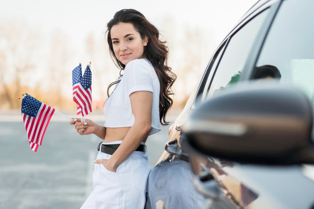 Chica morena con banderas de EEUU apoyada en un coche