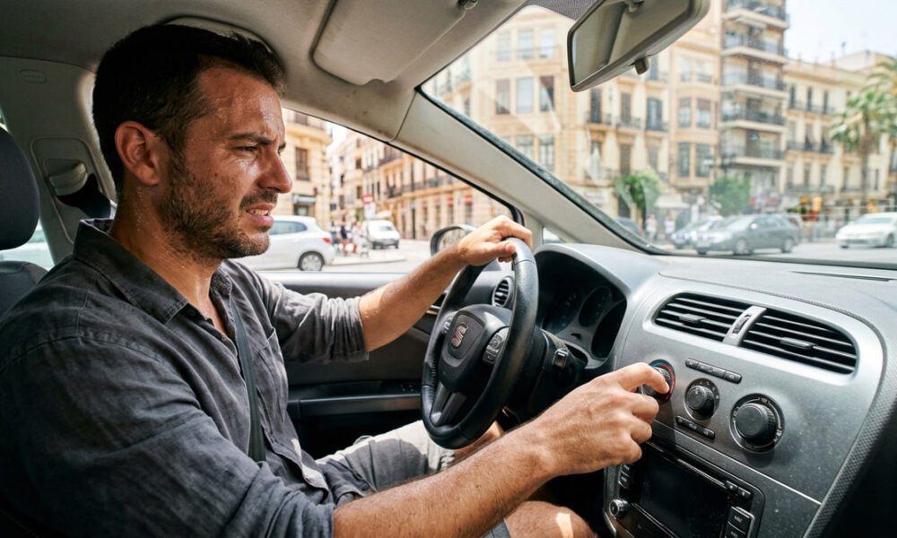 Conductor incómodo en pleno verano dentro del coche intentando ajustar el aire acondicionado sin éxito.