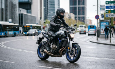 Imagen hiperrealista de un motorista con equipación completa conduciendo una motocicleta de media cilindrada en entorno urbano español, con asfalto húmedo y estilo editorial de revista del motor.