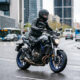 Imagen hiperrealista de un motorista con equipación completa conduciendo una motocicleta de media cilindrada en entorno urbano español, con asfalto húmedo y estilo editorial de revista del motor.